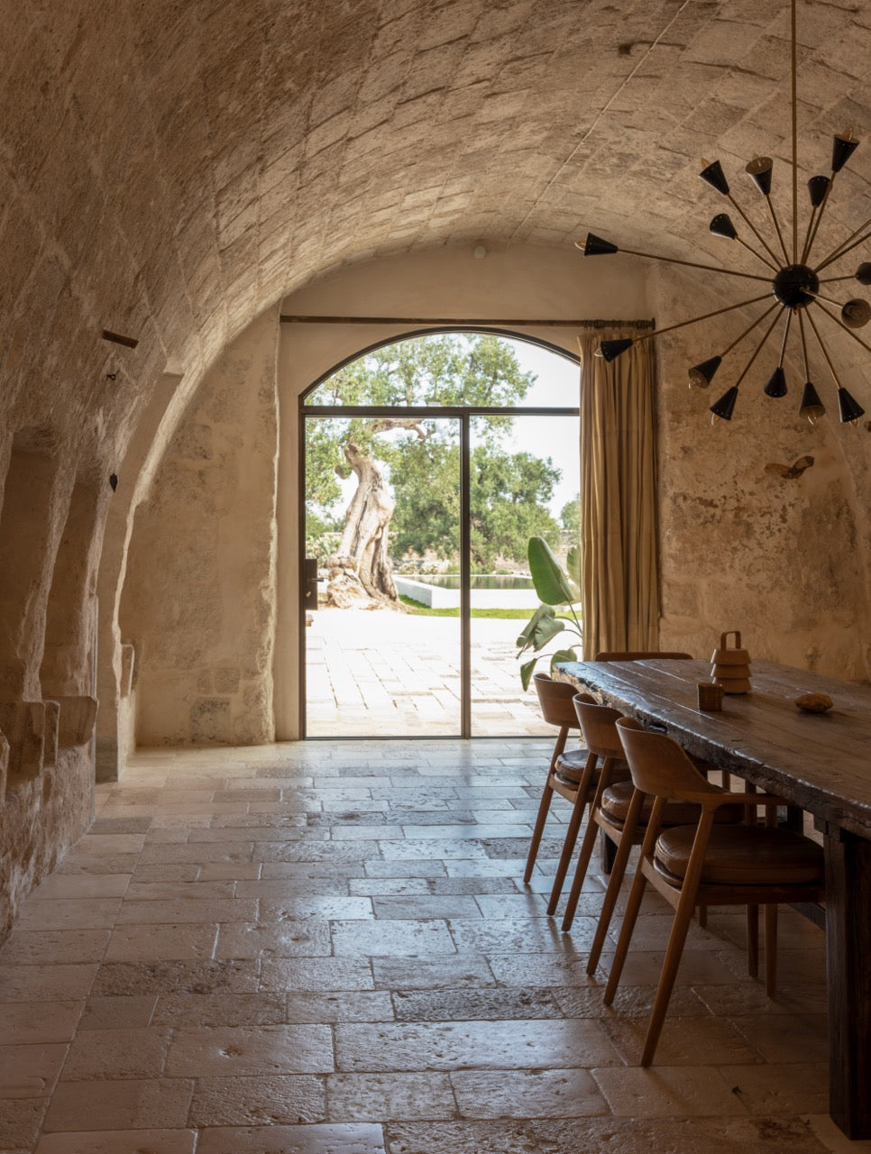 Vaulted dining room with wooden table opening onto pool and olive tree