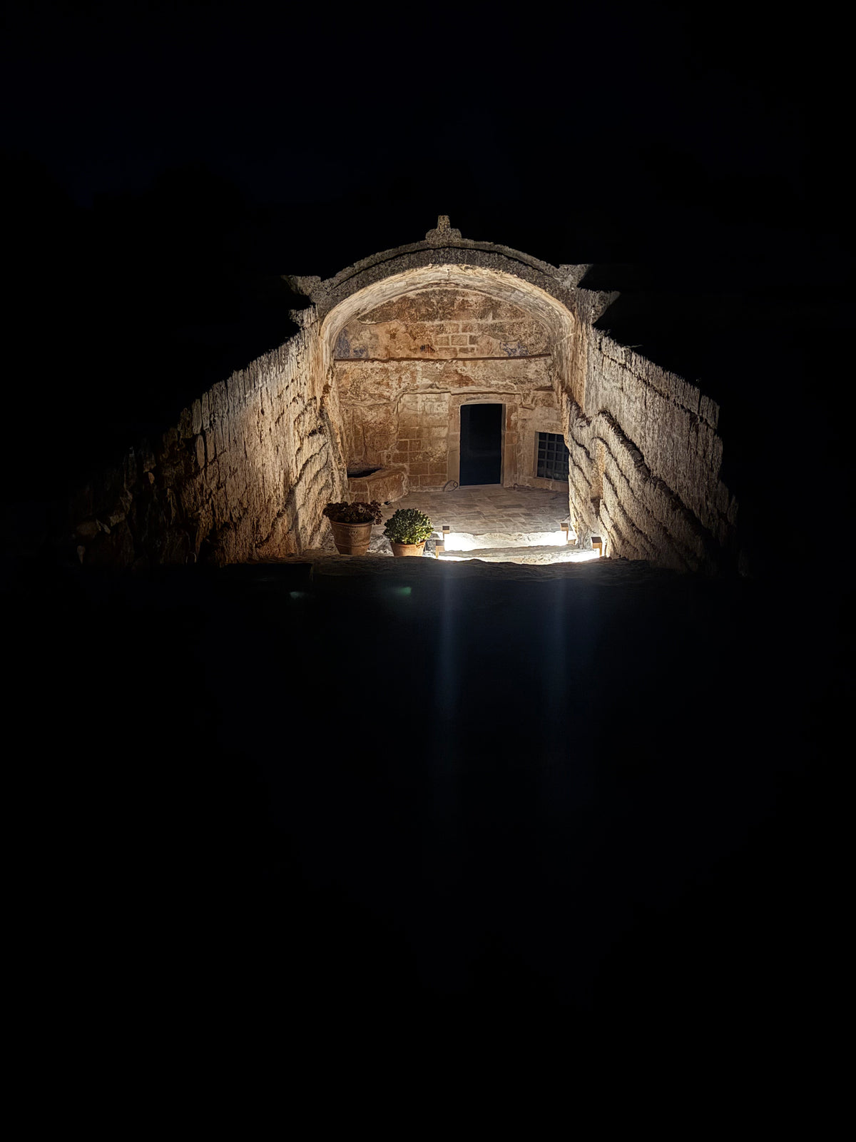 A dramatic night photograph of the cave entrance, dramatically lit from within to reveal textured stone walls and a doorway, set against a pitch-black background.