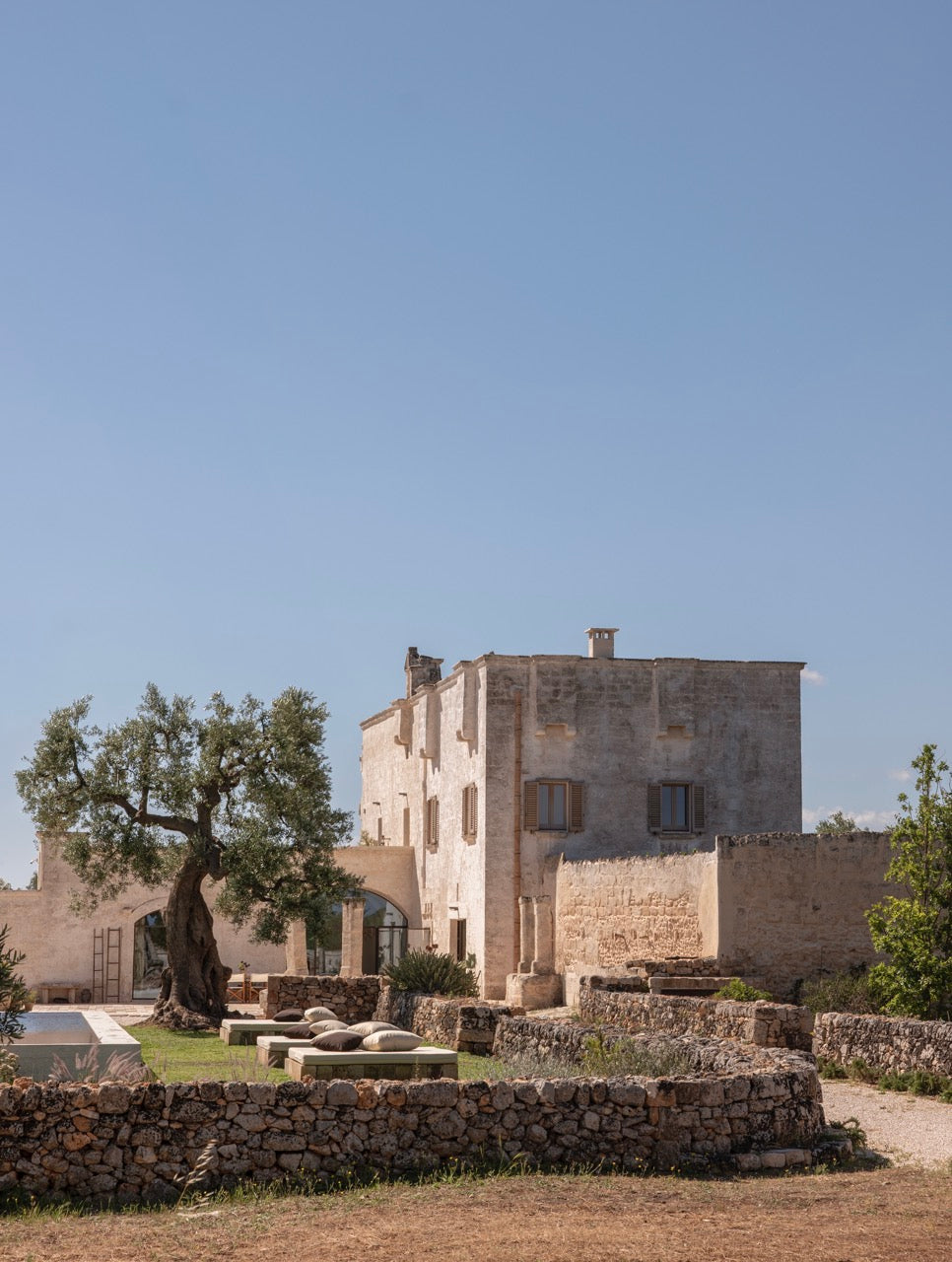 Stone farmhouse with pool and lounge seating beside olive tree